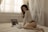 A person is kneeling on a bed, looking at documents in a softly lit room. A laptop is open beside them, and the ambiance is calm and intimate due to the natural light filtering through the blinds.