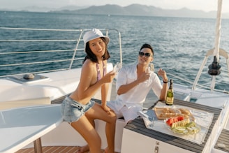 Two people are enjoying a sunny day on a yacht. One person is sitting on a table in denim shorts and a straw hat, holding a glass, while another person is dressed in a white outfit, wearing sunglasses, and gesturing with his hand. On the table, there is a spread of food including bread, fruit slices, and a bottle of champagne. The backdrop features a calm sea with mountains in the distance.