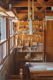 A cozy wooden workshop filled with various materials and crafted items. The space includes shelves displaying wooden crafts and tools, with sunlight streaming in through large windows on the left. Wind chimes and other decorative pieces hang from the ceiling.