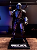 A detailed figure of a character in futuristic armor stands with a blaster in hand. The armor is metallic with a sleek design, and the character wears a helmet with a T-shaped visor. The figure is mounted on a display stand with the words 'Star Wars The Mandalorian' prominently displayed. The background is dimly lit, casting a dramatic shadow around the figure.