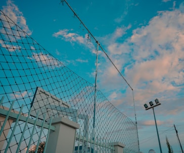 A tall chain-link fence topped with barbed wire stands against a backdrop of a blue sky scattered with fluffy, pink-tinged clouds. Nearby, stadium lights are seen, suggesting a sports field setting.