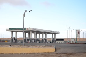 A petrol station in an empty, arid landscape, featuring multiple fuel pumps under a modern canopy. The station's branding is visible with signs in both English and Arabic. The environment appears deserted, with a clear blue sky and distant hills, creating a sense of isolation.