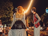 Two performers wearing striped costumes are standing on a bustling street at night. The woman on the left has a black lace accent on her back, while the man on the right wears a red sequined vest. The street is lit with string lights, and several people are visible in the background near trees and parked cars.