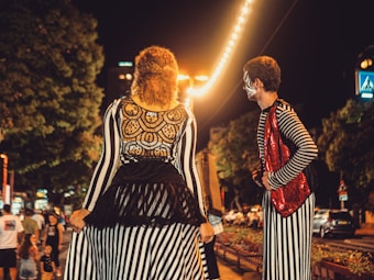 Two performers wearing striped costumes are standing on a bustling street at night. The woman on the left has a black lace accent on her back, while the man on the right wears a red sequined vest. The street is lit with string lights, and several people are visible in the background near trees and parked cars.