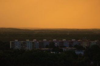A row of apartment buildings is silhouetted against a glowing, orange sky. The horizon is filled with dense forests and rolling hills, creating a tranquil and expansive atmosphere.