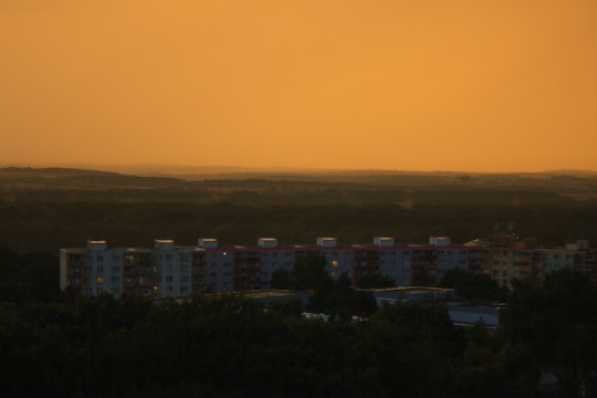 A row of apartment buildings is silhouetted against a glowing, orange sky. The horizon is filled with dense forests and rolling hills, creating a tranquil and expansive atmosphere.