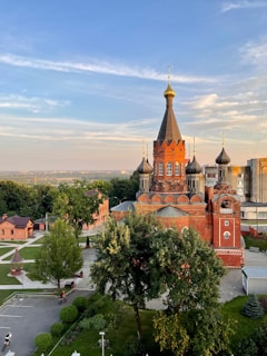 A red brick building with traditional Eastern European architecture is centered in a park-like setting. The building has ornate details and several domes topped with crosses. Surrounding the building are well-maintained trees and pathways. In the background, a cityscape is visible under a blue sky with light clouds. There are a few people visible near the building and on bicycles.
