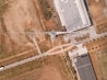 An aerial view of an industrial area featuring a large wind turbine situated adjacent to a facility with rooftop solar panels. Surrounding the structures, there are patches of grass and dirt with a network of roads and pathways visible. A few scattered trees line the streets and there are several parked cars near the building.