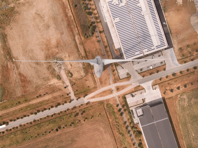 An aerial view of an industrial area featuring a large wind turbine situated adjacent to a facility with rooftop solar panels. Surrounding the structures, there are patches of grass and dirt with a network of roads and pathways visible. A few scattered trees line the streets and there are several parked cars near the building.