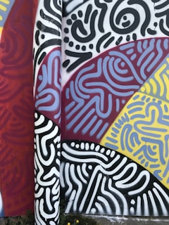 A vibrant graffiti mural featuring abstract patterns and shapes. The artwork includes a mix of swirling and linear designs in colors such as black, white, red, yellow, and blue. The overall composition is intricate, with each section creating a distinct visual texture.