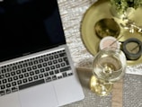 A laptop with a black screen is placed on a textured surface next to a glass of white wine on a golden tray. The tray also holds small bowls with condiments and a potted plant for decoration.