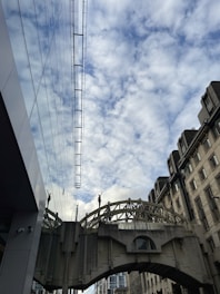 An architectural archway spans between two buildings, adorned with sculptures. The sky is filled with scattered clouds, and the modern building on one side reflects the structure opposite.