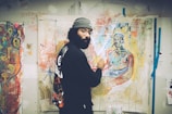 An artist is painting in a brightly colored studio filled with abstract artworks. The wall is covered with expressive and vibrant paintings. The artist, dressed in casual, artistic attire, focuses on a canvas depicting a figure.