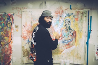 An artist is painting in a brightly colored studio filled with abstract artworks. The wall is covered with expressive and vibrant paintings. The artist, dressed in casual, artistic attire, focuses on a canvas depicting a figure.