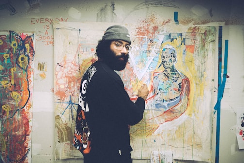 An artist is painting in a brightly colored studio filled with abstract artworks. The wall is covered with expressive and vibrant paintings. The artist, dressed in casual, artistic attire, focuses on a canvas depicting a figure.