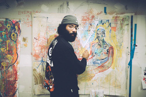An artist painting a bold, expressive mural in a sunlit studio filled with art supplies.