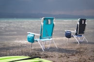 Two folding beach chairs are placed in shallow water near the shore. The chairs are blue and turquoise with storage bags attached. A kayak or paddleboard is partially visible in the foreground. The background showcases calm waters extending towards distant mountains under a cloudy sky.