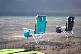 Two folding beach chairs are placed in shallow water near the shore. The chairs are blue and turquoise with storage bags attached. A kayak or paddleboard is partially visible in the foreground. The background showcases calm waters extending towards distant mountains under a cloudy sky.