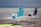 Two folding beach chairs are placed in shallow water near the shore. The chairs are blue and turquoise with storage bags attached. A kayak or paddleboard is partially visible in the foreground. The background showcases calm waters extending towards distant mountains under a cloudy sky.