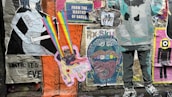 A collage of colorful street art posters on a wall, featuring a variety of graphic designs and styles. One poster has a cartoonish character emitting rainbow beams from its eyes, while another displays text about 'acid tabs'. There are printed images of people, abstract patterns, and cutouts of human figures.