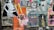 A collage of colorful street art posters on a wall, featuring a variety of graphic designs and styles. One poster has a cartoonish character emitting rainbow beams from its eyes, while another displays text about 'acid tabs'. There are printed images of people, abstract patterns, and cutouts of human figures.