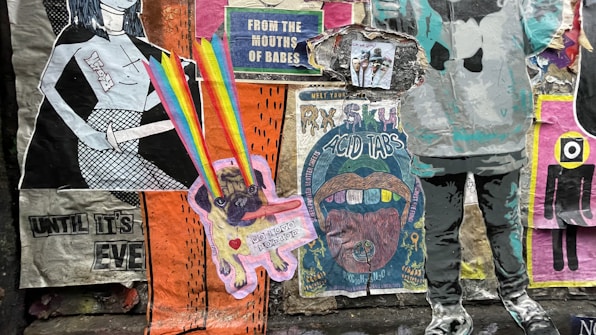 A collage of colorful street art posters on a wall, featuring a variety of graphic designs and styles. One poster has a cartoonish character emitting rainbow beams from its eyes, while another displays text about 'acid tabs'. There are printed images of people, abstract patterns, and cutouts of human figures.