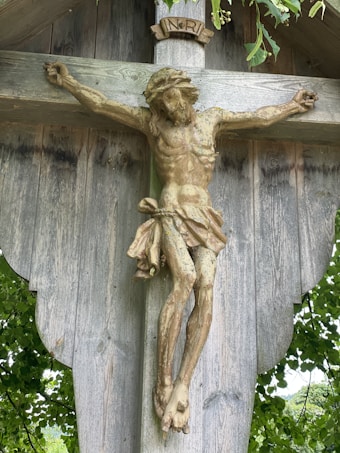 A wooden crucifix depicting Jesus Christ with an inscription above his head. The cross is weathered and surrounded by lush green leaves providing a natural backdrop.