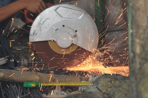 Sparks fly as a circular saw cuts through a piece of material. A person's hand is visible, guiding the tool, while various other objects surround the work area, including a measuring tape and materials like wood and metal.