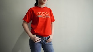A person wearing a bright red Gucci t-shirt paired with blue jeans stands against a plain wall. Their hands are placed on their hips, and their head is partially cropped out of the frame.
