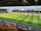 A sports stadium with a well-maintained grass field is depicted. Players are spread across the field, engaging in warm-up activities. The stands are partially filled with spectators, and the stadium is surrounded by urban buildings under a partly cloudy sky.