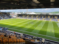 A sports stadium with a well-maintained grass field is depicted. Players are spread across the field, engaging in warm-up activities. The stands are partially filled with spectators, and the stadium is surrounded by urban buildings under a partly cloudy sky.