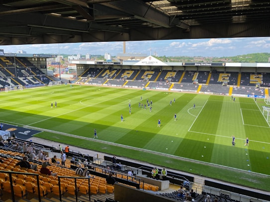 A sports stadium with a well-maintained grass field is depicted. Players are spread across the field, engaging in warm-up activities. The stands are partially filled with spectators, and the stadium is surrounded by urban buildings under a partly cloudy sky.