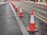 A row of orange and white traffic cones is lined up along a wet street, guiding vehicles away from the tram tracks. Additional traffic cones and pink barriers are visible in the background, indicating a construction or maintenance zone. The pavement appears to be slightly wet, suggesting recent rainfall.