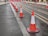 A row of orange and white traffic cones is lined up along a wet street, guiding vehicles away from the tram tracks. Additional traffic cones and pink barriers are visible in the background, indicating a construction or maintenance zone. The pavement appears to be slightly wet, suggesting recent rainfall.