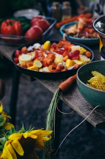 A rustic table is adorned with an assortment of colorful vegetables and fresh produce. There are slices of various vegetables like corn, carrots, and squash in a bowl. Nearby, bright sunflowers add a touch of nature, while whole red bell peppers are visible in the background. A lone carrot with greens attached lies across the table, emphasizing a harvest theme.