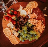 A wooden platter loaded with assorted snacks, including fresh strawberries, green grapes, a variety of crackers, soft cheese, nuts, and some type of dried fruit. The arrangement is visually appealing, inviting, and casual, with some crumbs and a spreader lying nearby, suggesting the platter is in use. Wine glasses are partially visible around the platter.