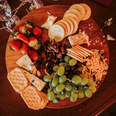 A wooden platter loaded with assorted snacks, including fresh strawberries, green grapes, a variety of crackers, soft cheese, nuts, and some type of dried fruit. The arrangement is visually appealing, inviting, and casual, with some crumbs and a spreader lying nearby, suggesting the platter is in use. Wine glasses are partially visible around the platter.
