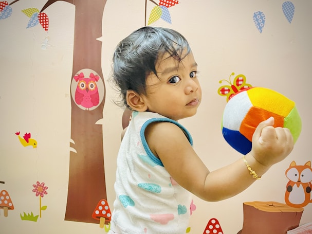 A child is holding a colorful soft ball while looking back. The room has playful stickers on the wall, including a tree, owls, birds, and mushrooms, creating a whimsical atmosphere.
