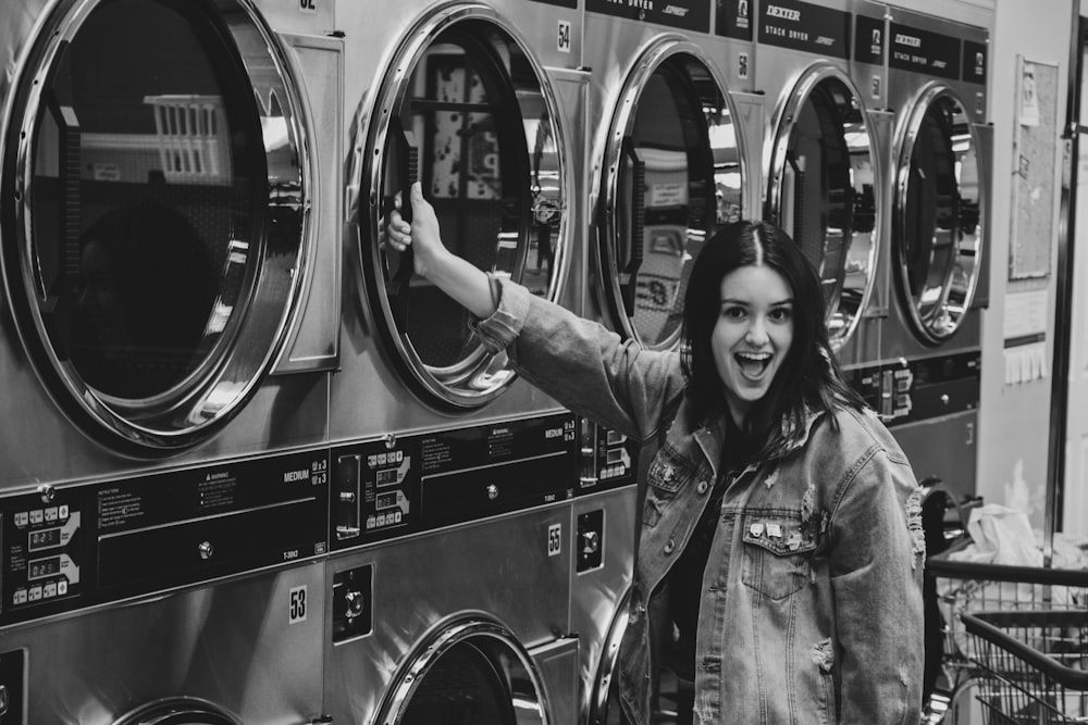 grayscale photo of woman in button up shirt standing beside front load washing machine