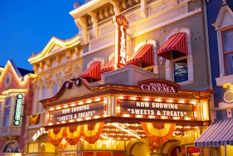 An ornate cinema façade with bright lights and signs. The marquee displays 'Main St. Cinema' and 'Now Showing Sweets & Treats.' Red and white striped awnings adorn the windows above, and the building features a blend of classic architectural elements and festive decorations. The overall ambiance suggests a vintage or historic setting.