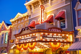 An ornate cinema fa&ccedil;ade with bright lights and signs. The marquee displays 'Main St. Cinema' and 'Now Showing Sweets & Treats.' Red and white striped awnings adorn the windows above, and the building features a blend of classic architectural elements and festive decorations. The overall ambiance suggests a vintage or historic setting.