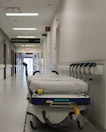 A hospital hallway is depicted with a gurney positioned in the foreground. The corridor is clean and well-lit, with signs indicating different departments such as 'GASTROENTEROLOGIE.' Several closed doors and white walls line the hallway, creating a sterile and orderly environment.