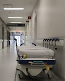 A hospital hallway is depicted with a gurney positioned in the foreground. The corridor is clean and well-lit, with signs indicating different departments such as 'GASTROENTEROLOGIE.' Several closed doors and white walls line the hallway, creating a sterile and orderly environment.