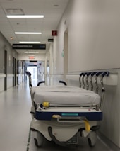A hospital hallway is depicted with a gurney positioned in the foreground. The corridor is clean and well-lit, with signs indicating different departments such as 'GASTROENTEROLOGIE.' Several closed doors and white walls line the hallway, creating a sterile and orderly environment.
