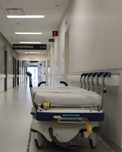 A hospital hallway is depicted with a gurney positioned in the foreground. The corridor is clean and well-lit, with signs indicating different departments such as 'GASTROENTEROLOGIE.' Several closed doors and white walls line the hallway, creating a sterile and orderly environment.