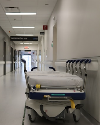 A hospital hallway is depicted with a gurney positioned in the foreground. The corridor is clean and well-lit, with signs indicating different departments such as 'GASTROENTEROLOGIE.' Several closed doors and white walls line the hallway, creating a sterile and orderly environment.