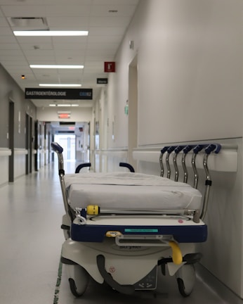 A hospital hallway is depicted with a gurney positioned in the foreground. The corridor is clean and well-lit, with signs indicating different departments such as 'GASTROENTEROLOGIE.' Several closed doors and white walls line the hallway, creating a sterile and orderly environment.