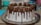 A beautifully decorated chocolate cake with a white frosting base. The cake has chocolate ganache dripping down the sides and is topped with chocolate truffles, each sprinkled with small gold beads.
