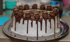 A beautifully decorated chocolate cake with a white frosting base. The cake has chocolate ganache dripping down the sides and is topped with chocolate truffles, each sprinkled with small gold beads.