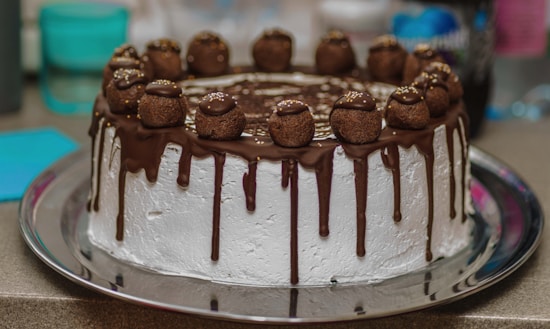 A beautifully decorated chocolate cake with a white frosting base. The cake has chocolate ganache dripping down the sides and is topped with chocolate truffles, each sprinkled with small gold beads.